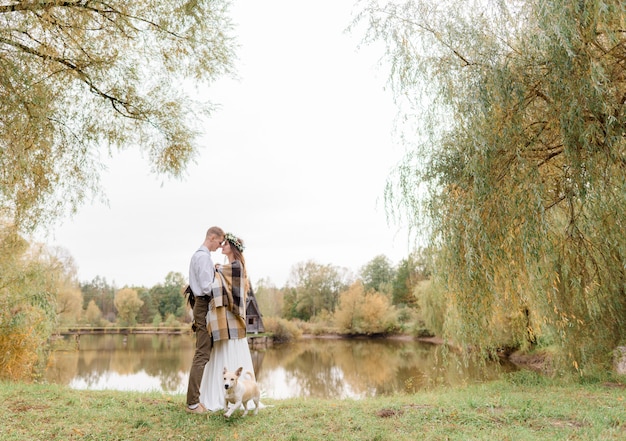 Tierna pareja de enamorados en el parque de otoño con un perro está de pie casi besándose cerca del lago