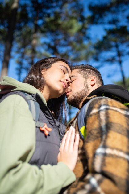 Tierna pareja de jóvenes excursionistas. Hombre caucásico con barba y mujer con gorra con grandes mochilas tocando caricias tiernamente en el bosque. Hobby, naturaleza, concepto de amor.