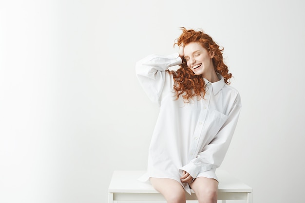 Tierna pelirroja joven en camisa sentada en la mesa riendo con los ojos cerrados corrigiendo el cabello.