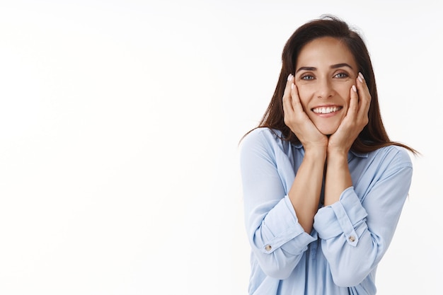 Tierna romántica alegre morena mujer de ojos azules con tatuajes, sintiendo felicidad y amor, suspirando románticamente, sonriendo ampliamente, mirando la admiración frontal, tocando las mejillas ruborizándose complacido