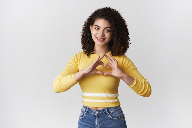 Tierna romántica joven cariñosa novia de pelo rizado expresar amor pasión simpatía y positividad mostrar corazón signo pecho sonriendo felizmente confesando sentimientos reconfortantes de pie fondo blanco