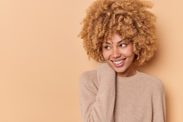 La tierna y sonriente mujer de pelo rizado mantiene la mano cerca del cuello y se ve alegremente lejos de estar de buen humor viste un jersey de manga larga aislado sobre fondo beige con espacio de copia para su anuncio