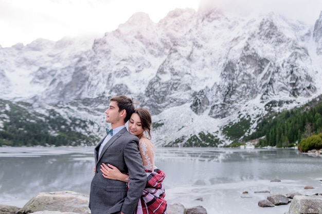 Tierna sonrisa pareja en traje de novia está de pie delante del hermoso paisaje de montaña de invierno