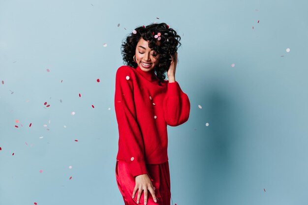 Tímida chica de raza mixta de pie bajo confeti Mujer joven rizada en suéter rojo posando con una sonrisa sincera