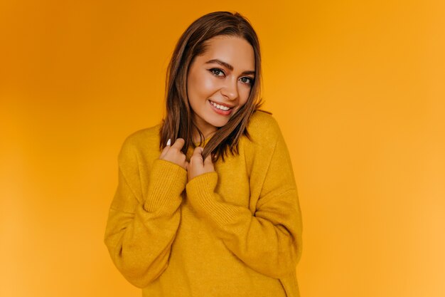 Tímida mujer europea posando con una sonrisa en la pared naranja. Chica atractiva en suéter amarillo divirtiéndose.