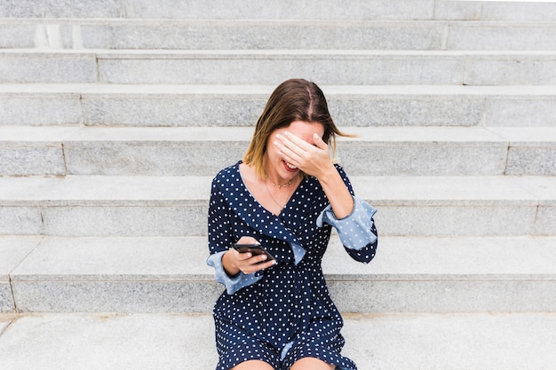 Foto gratuita tímida mujer joven sentada en la escalera con smartphone