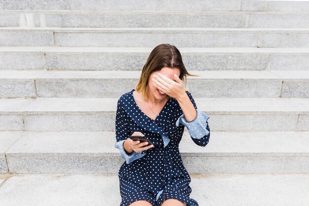 Tímida mujer joven sentada en la escalera con smartphone