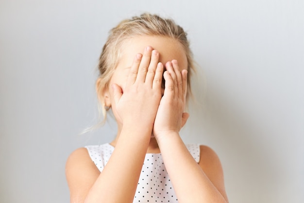 Tímida niña tímida que cubre la cara, sintiendo miedo. Niña avergonzada posando aislada con las manos en los ojos, llorando, sintiéndose avergonzada porque la madre la regaña. Bebé jugando al escondite