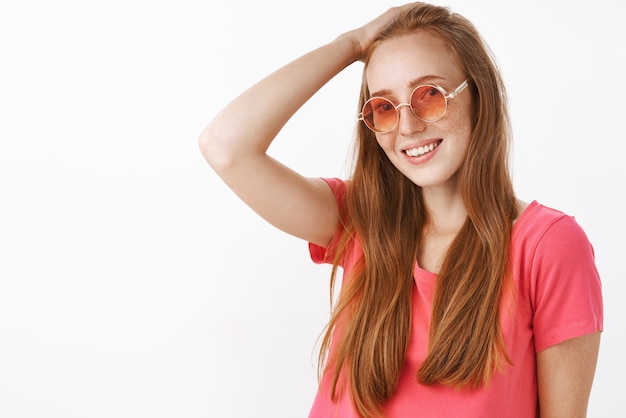 Tímida y tierna dama encantadora pelirroja con pecas en gafas de sol de moda y blusa rosa peinándose con la mano en la cabeza y sonriendo posando amigable