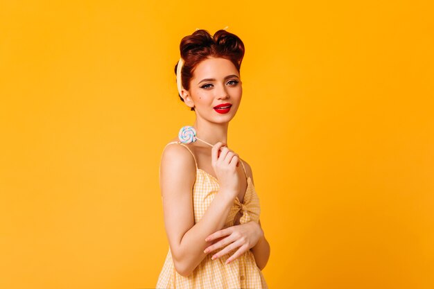 Tímido jengibre joven sosteniendo piruleta. Foto de estudio de glamorosa chica pinup sosteniendo dulces en el espacio amarillo.