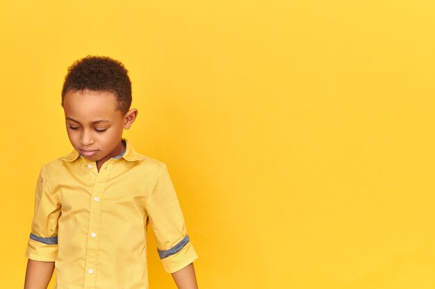 Tímido niño africano tímido que se avergüenza mirando hacia abajo sintiéndose avergonzado de mal comportamiento aislado en la pared amarilla en blanco