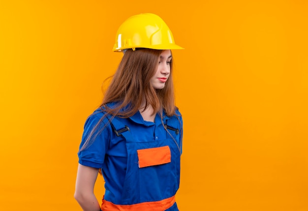 Tímido trabajador constructor joven en uniforme de construcción y casco de seguridad mirando con expresión triste