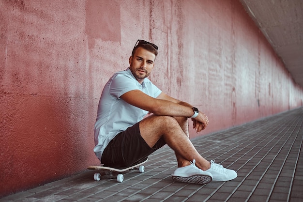 Un tipo apuesto y de moda vestido con una camisa blanca y pantalones cortos sentados en una patineta debajo del puente, mirando una cámara.