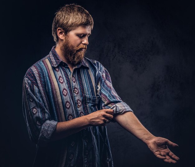 Un tipo barbudo pelirrojo vestido con una camisa pasada de moda se sube la manga. Aislado en un fondo oscuro.