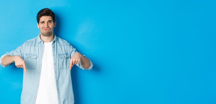 Foto gratuito un tipo decepcionado con ropa informal apuntando con el dedo hacia abajo con una mueca dudosa de pie contra el azul