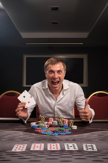 Foto gratuita un tipo emocional feliz con una camisa blanca está jugando al póquer sentado en la mesa del casino. se regocija en ganar mostrando dos ases. juegos de azar por dinero. juegos de fortuna.