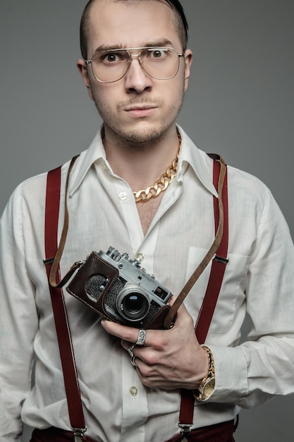 Un tipo extraño con gafas de sol y camisa blanca sostiene una cámara de cine sobre un fondo gris.