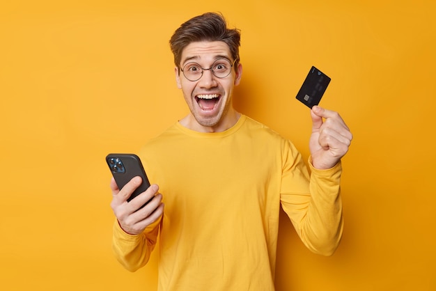 Un tipo guapo y alegre hace que la banca móvil sea segura a través de una aplicación celular, tiene una tarjeta de crédito, proporciona pagos por Internet, usa anteojos redondos y un saltador casual aislado sobre un fondo amarillo