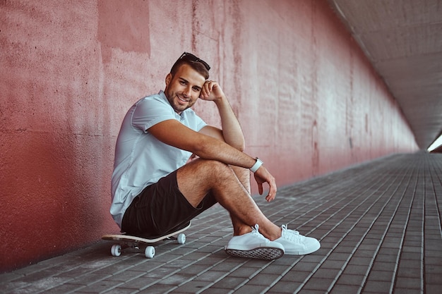 Un tipo guapo y sonriente vestido con una camisa blanca y pantalones cortos sentados en una patineta debajo del puente, mirando una cámara.