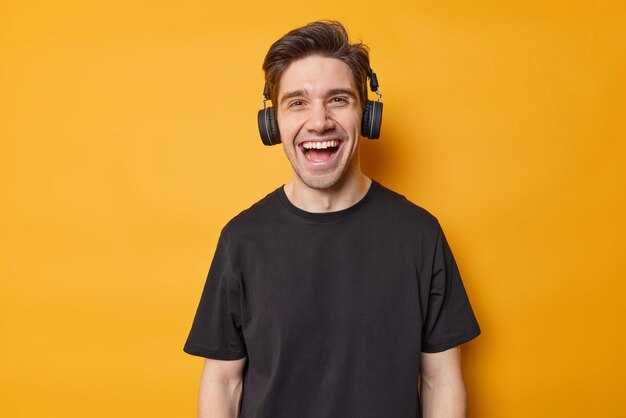 El tipo hipster feliz se ríe alegremente tiene un estado de ánimo optimista escucha su música favorita a través de auriculares estéreo usa una camiseta negra casual aislada sobre un fondo amarillo vívido Concepto de hobby y estilo de vida de la gente