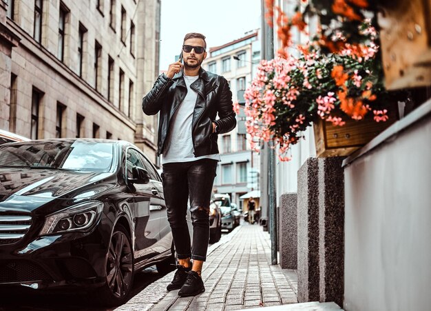 Un tipo de moda con gafas de sol vestido con una chaqueta de cuero negro y jeans habla con un smartphone mientras está parado cerca de un camión de lujo en una vieja calle de Europa.