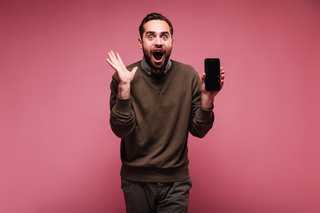 Un tipo optimista con suéter marrón mira felizmente a la cámara y sostiene el teléfono