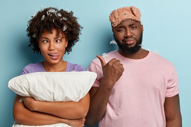 Un tipo de piel oscura disgustado señala a una divertida mujer afroamericana que cruza los ojos y saca la lengua, sostiene la almohada blanca y se divierte antes de irse a la cama. Pareja tiene régimen tardío, necesita tiempo de descanso