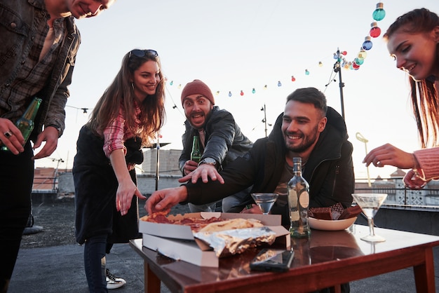 Foto gratuita el tipo del sombrero rojo hace una mueca de hambre y trata de alcanzar ese producto fresco. comiendo pizza en la fiesta de la azotea. los buenos amigos tienen un fin de semana con deliciosa comida y alcohol.