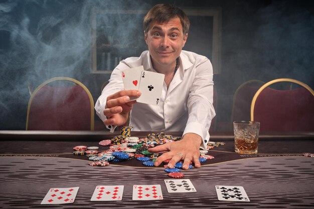 Un tipo sonriente y encantado con una camisa blanca está jugando al póquer sentado en la mesa del casino con humo. Se regocija en ganar mostrando dos ases a la cámara. Juegos de azar por dinero. Juegos de fortuna.