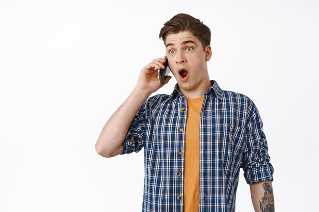 Un tipo sorprendido recibe noticias increíbles en una llamada telefónica, llama a alguien y se queda sin aliento asombrado, asombrado por las noticias, conversando en un teléfono inteligente, de pie contra un fondo blanco