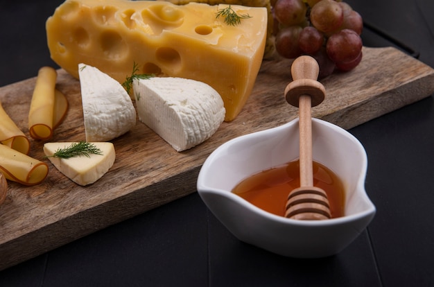 Tipo de vista lateral de queso en un soporte con uvas y miel sobre un fondo negro