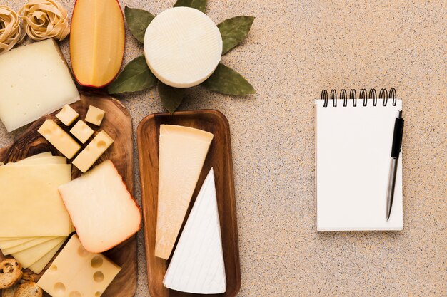 Tipos sanos de quesos en la placa de madera con la libreta y la pluma blancas en blanco