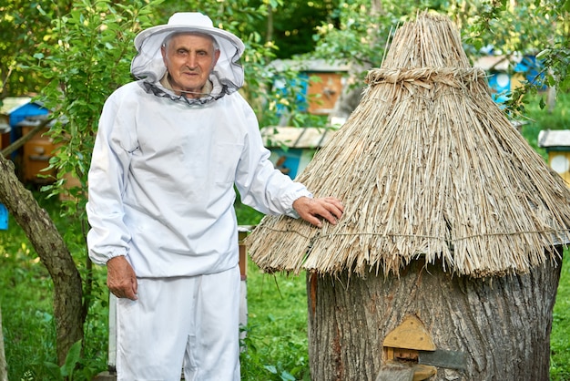 Tirado de un apicultor masculino mayor que lleva el traje de la apicultura que presenta en su colmenar cerca del concepto de la jubilación del estilo de vida de la profesión de la agricultura del copyspace de la colmena.