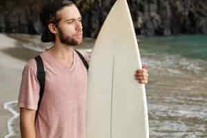 Foto gratuita tiro al aire libre del atractivo surfista barbudo sosteniendo su tabla de surf blanca de pie en la playa y mirando al océano, decidiéndose antes del concurso de surf