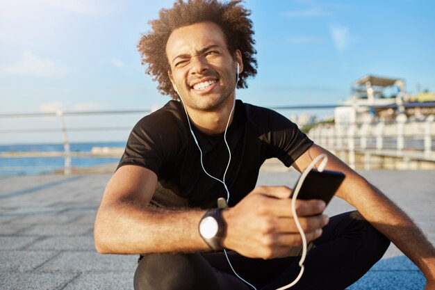 Tiro al aire libre de deportista afroamericano con peinado tupido entrecerrar los ojos debido al sol en ropa deportiva negra. Atleta masculino de piel oscura sentado con las piernas cruzadas sosteniendo en su mano teléfono celular