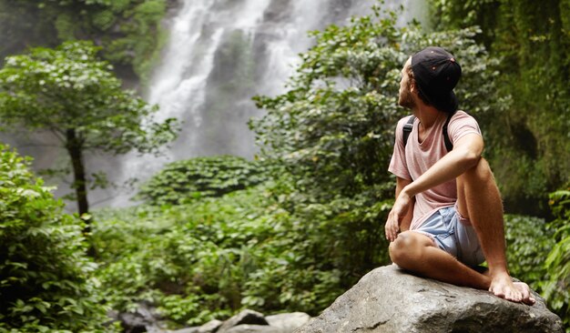 Tiro al aire libre del excursionista caucásico joven y elegante sentado descalzo sobre una gran roca y mirando por encima del hombro a la hermosa cascada