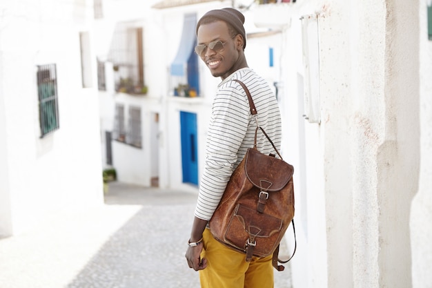 Tiro al aire libre de guapo feliz joven viajero masculino afroamericano con mochila de cuero de pie en el muro de hormigón en la calle estrecha mientras visita la ciudad turística durante sus vacaciones de verano