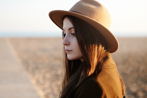 Tiro al aire libre de la hermosa joven europea con cabello largo y oscuro con sombrero mientras caminaba por la playa de la ciudad, sintiéndose triste y sola