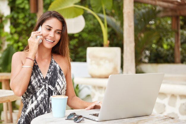 Tiro al aire libre de hermosa mujer morena con expresión alegre, tiene conversación móvil con un amigo, revisa el correo electrónico en línea en la computadora portátil