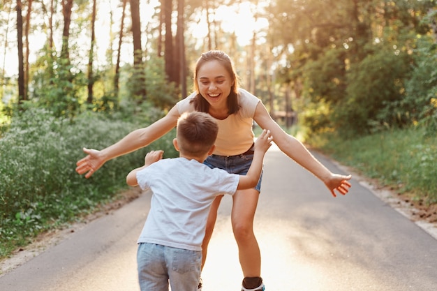 Tiro al aire libre de hermosa mujer con sonrisa dentuda y expresión feliz positiva atrapando a su hijo con los brazos abiertos mientras patinan juntos en el parque de verano, estilo de vida activo y saludable.