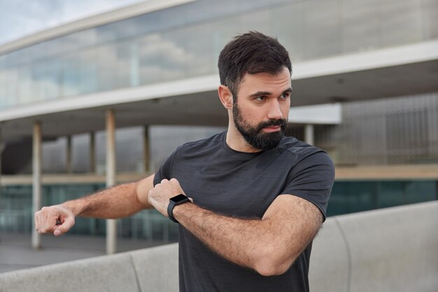 Tiro al aire libre de hombre barbudo motivado estira los brazos se calienta antes de que el entrenamiento use camiseta negra casual smartwacth concentrada en poses de distancia contra borrosa. Concepto de deporte.