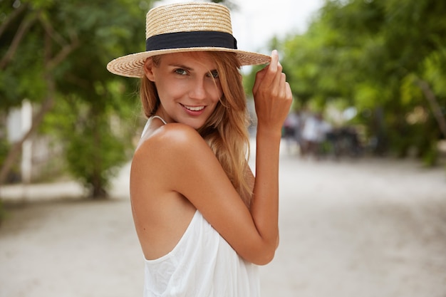 Tiro al aire libre de mujer de aspecto agradable con piel bronceada y saludable, vestida con vestido blanco y sombrero de verano, posa en el parque con expresión de confianza y satisfacción