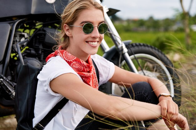 Tiro al aire libre de mujer hermosa activa positiva en tonos de moda, usa pañuelo rojo en el cuello, pasa tiempo libre al aire libre como paseos en motocicleta, tiene un viaje extremo. Concepto de personas, descanso y pasatiempo.