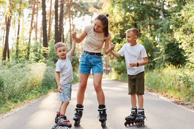 Tiro al aire libre de mujer joven atractiva con camiseta beige y pantalones vaqueros cortos patinar con niños, madre y niños expresando emociones positivas, pasatiempo en el parque de verano.