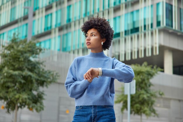 Tiro al aire libre de mujer joven pensativa con cabello rizado comprueba el tiempo en el reloj de pulsera espera a que alguien en la calle concentrado en la distancia use un jersey casual y jeans se encuentra en un entorno urbano.