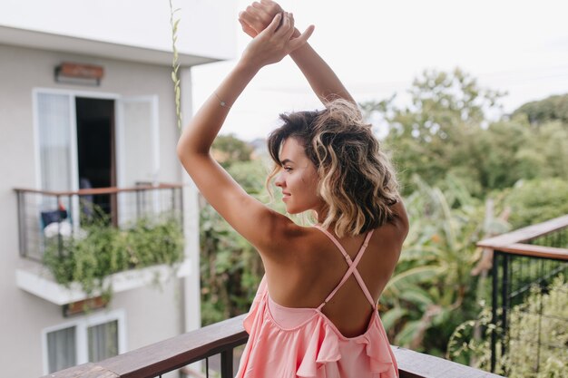 Tiro al aire libre de la parte posterior de la mujer bronceada con cabello ondulado rubio. Hermosa modelo femenina agitando las manos mientras posa en la terraza del hotel.