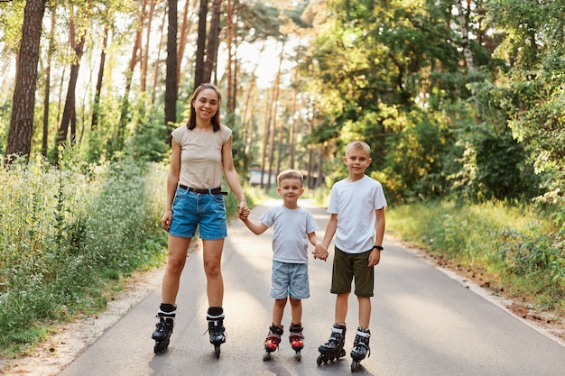 Tiro al aire libre de sonriente mujer atractiva con sus pequeños hijos de pie en la carretera en el parque de verano y tomados de la mano, familia patinar juntos, divertirse, pasatiempo activo.