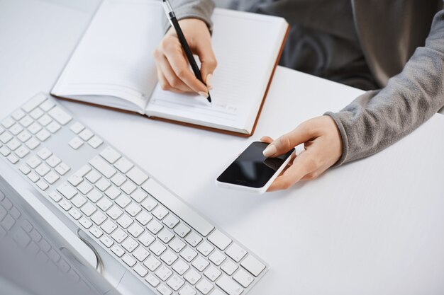 Tiro de alto ángulo de manos de mujer trabajando con gadgets. Captura recortada del teléfono inteligente femenino moderno que sostiene mientras escribe el plan en el cuaderno, sentado cerca del teclado y la computadora, teniendo dificultades en la oficina