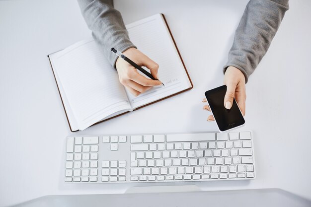 Tiro de alto ángulo de mujer trabajadora ocupada con smartphone. Captura recortada de manos de mujer haciendo notas en el planificador y sentado cerca del teclado y la computadora, trabajando y trabajando con compañeros de equipo.