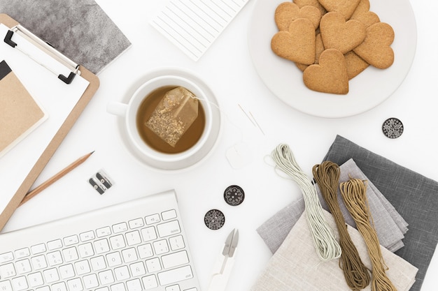 Tiro de alto ángulo de un teclado, una taza de té y galletas, algunos hilos y papeles sobre una superficie blanca